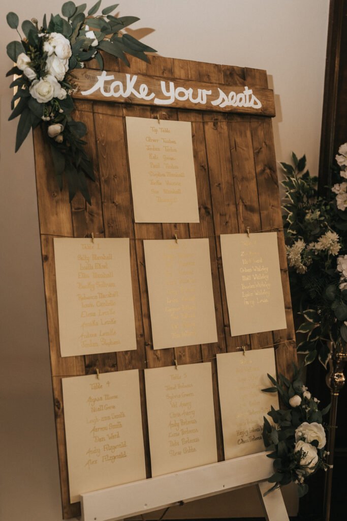 A rustic wooden seating chart display with “take your seats” written in white script at the top. Nine beige cards with gold calligraphy list guest names. Greenery and white flowers decorate the upper left and lower right corners. © Aimee Lince Photography, Lincolnshire photographer