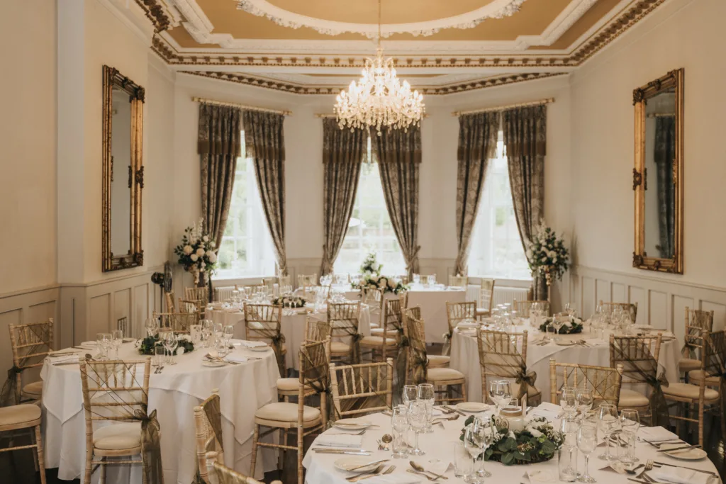 Elegant banqueting hall with round tables covered in white cloths, gold chairs, floral centrepieces, and place settings. Large windows with ornate curtains, gold-framed mirrors on the walls, and a sparkling chandelier hanging from an intricate ceiling. © Aimee Lince Photography, Lincolnshire photographer