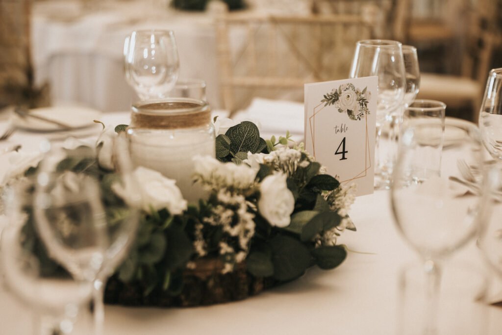 A close-up of an elegant wedding table centrepiece featuring white flowers, greenery, a white candle in a glass jar, and a "Table 4" card with floral designs, all arranged on a wooden base. Surrounding glasses and plates complete the refined setting. © Aimee Lince Photography, Lincolnshire photographer