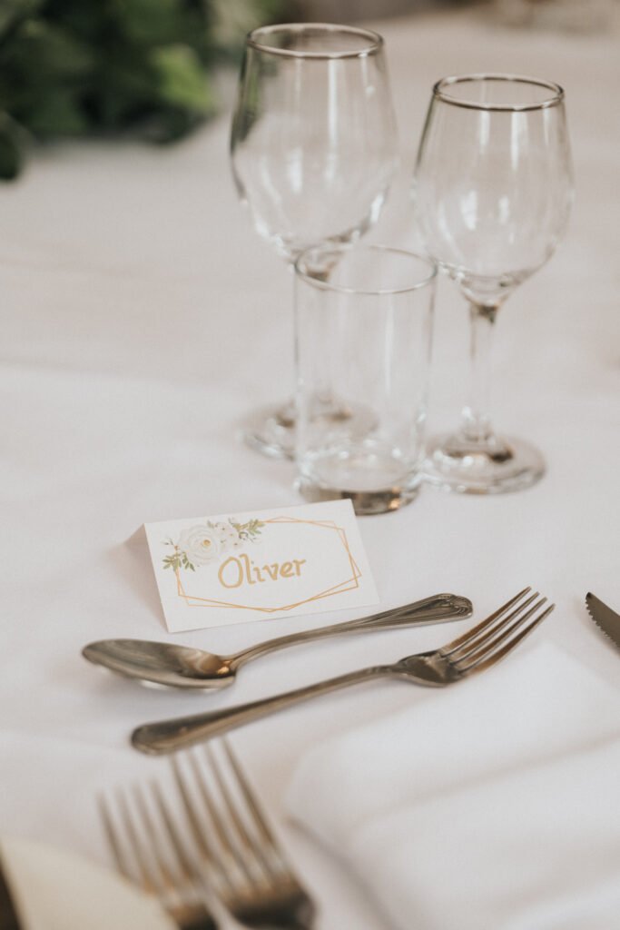A formal table setting with two forks, two spoons, and a knife on a white tablecloth, next to a folded serviette. Three glasses (two wine, one water) stand behind a name card labelled "Oliver" with a floral design. Greenery is blurred in the background. © Aimee Lince Photography, Lincolnshire photographer