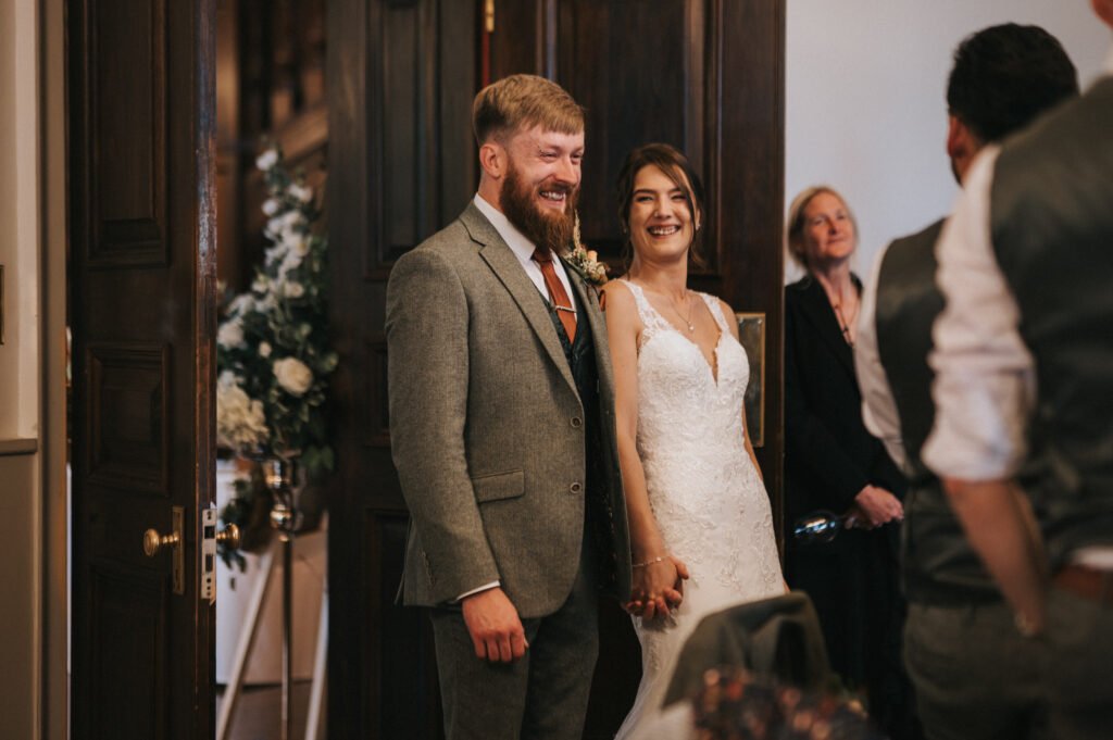 A smiling bride in a white lace gown and a groom in a grey suit stand holding hands in a warmly lit room, looking happy. They are surrounded by wooden doors, floral arrangements, and guests, suggesting a wedding ceremony or celebration. © Aimee Lince Photography, Lincolnshire photographer