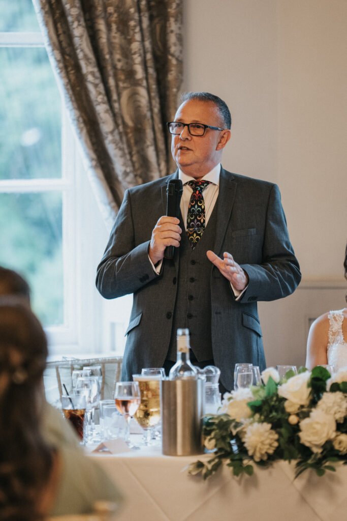 A middle-aged man in a grey suit and patterned tie stands at a banquet table, holding a microphone and speaking. He is near a window with patterned curtains; floral centrepieces, glasses, and a wine bottle are on the table in front of him. © Aimee Lince Photography, Lincolnshire photographer