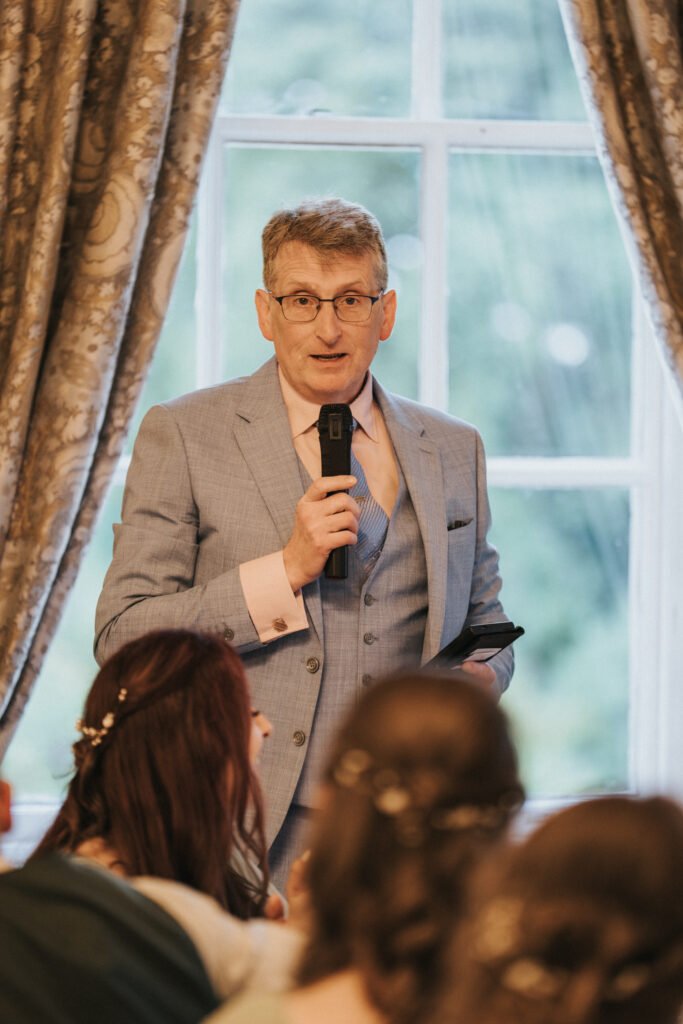 An older man in a light blue suit, pink shirt, and glasses holds a microphone and speaks indoors. He stands in front of large windows framed by ornate curtains, with blurred people seated in the foreground, likely at a formal event. © Aimee Lince Photography, Lincolnshire photographer
