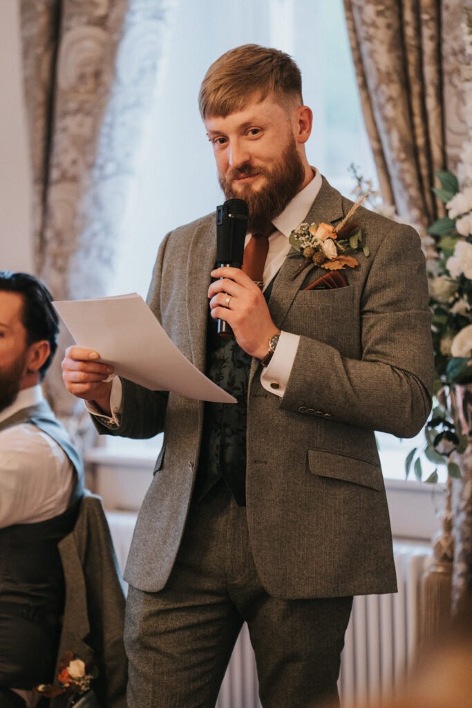 A man in a grey suit with a buttonhole holds a microphone and a sheet of paper, speaking at an indoor event. He has a short beard and styled hair. Elegant curtains and floral arrangements are visible in the background. © Aimee Lince Photography, Lincolnshire photographer