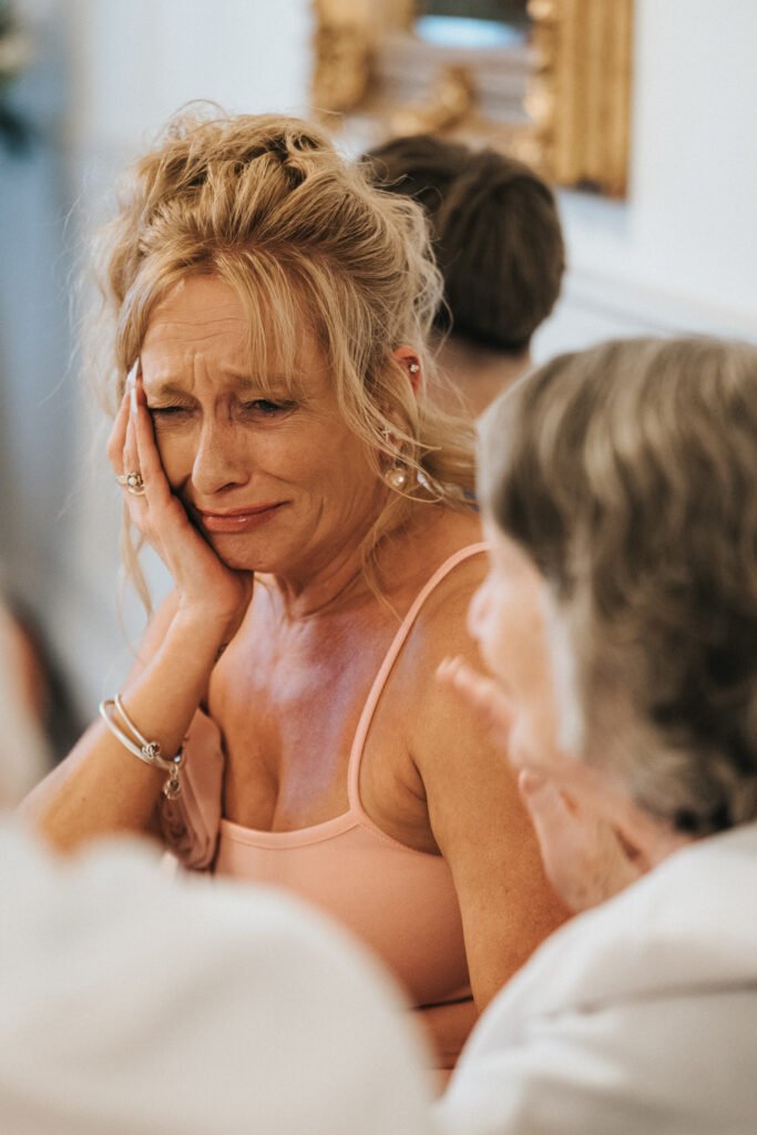 A woman with blonde hair in an updo and wearing a pink dress sits indoors, touching her face and appearing emotional or tearful. She is surrounded by other people, with one older woman in the foreground, slightly out of focus. © Aimee Lince Photography, Lincolnshire photographer