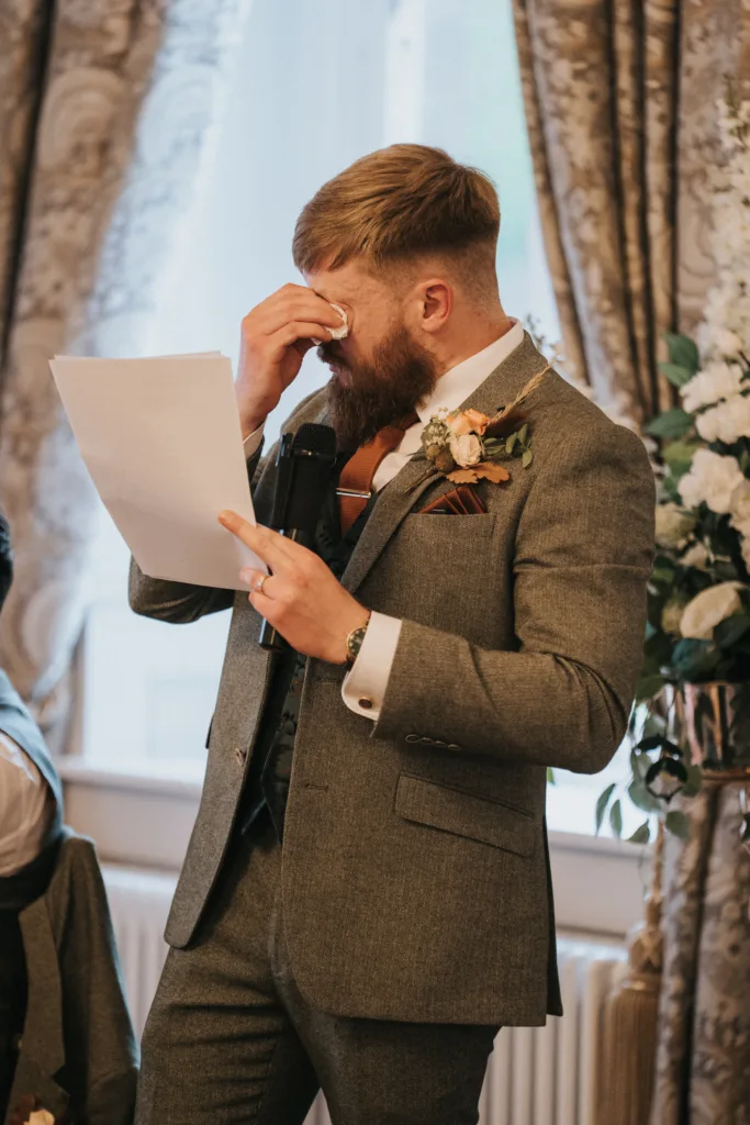 A bearded man in a grey suit wipes his eyes with a tissue while holding a piece of paper and a microphone. He stands in an ornate room with floral arrangements and patterned curtains, appearing emotional, likely whilst giving a speech. © Aimee Lince Photography, Lincolnshire photographer