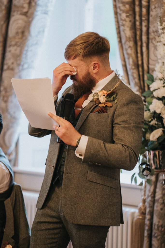 A bearded man in a grey suit wipes his eyes with a tissue while holding a piece of paper and a microphone. He stands in an ornate room with floral arrangements and patterned curtains, appearing emotional, likely whilst giving a speech. © Aimee Lince Photography, Lincolnshire photographer