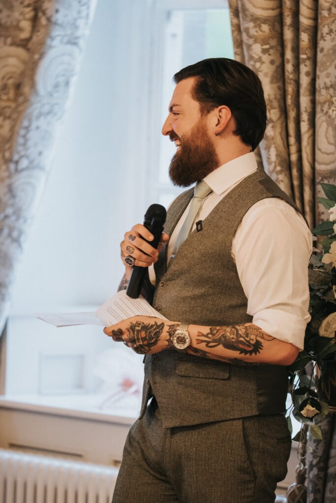 A bearded man with tattoos, wearing a grey waistcoat, white shirt, and light green tie, smiles while holding a microphone and cue cards. He stands indoors by a window with patterned curtains, appearing to give a speech at a formal event. © Aimee Lince Photography, Lincolnshire photographer