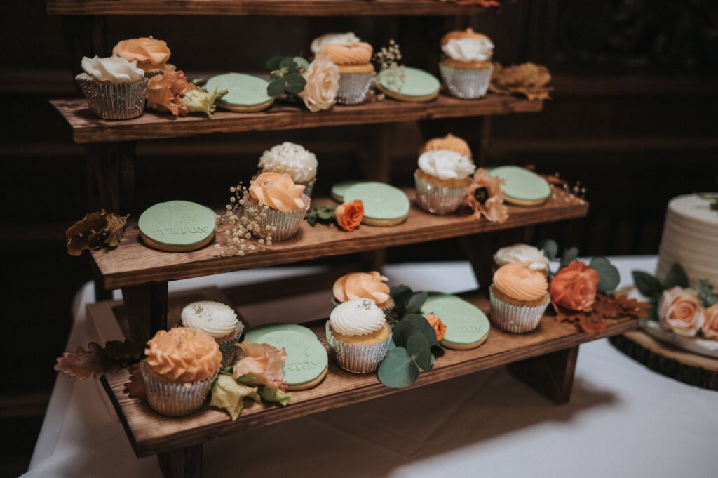 A three-tier wooden stand holds cupcakes with peach and white icing, green biscuits, and peach and cream flowers with foliage. The display sits on a white tablecloth, creating a rustic, elegant dessert arrangement. © Aimee Lince Photography, Lincolnshire photographer