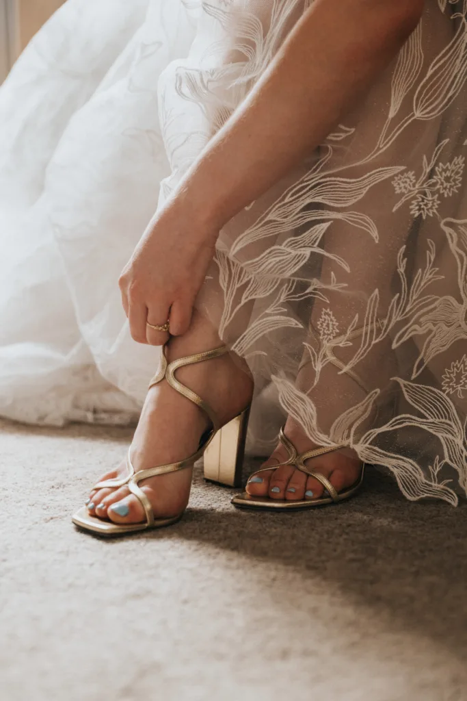 A woman in a delicate, floral-patterned dress adjusts a gold strappy sandal on her foot, with her toes painted light blue, as natural light illuminates the scene. © Aimee Lince Photography, Wedding photographer in Lincolnshire