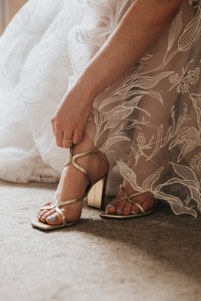 A woman in a delicate, floral-patterned dress adjusts a gold strappy sandal on her foot, with her toes painted light blue, as natural light illuminates the scene. © Aimee Lince Photography, Wedding photographer in Lincolnshire