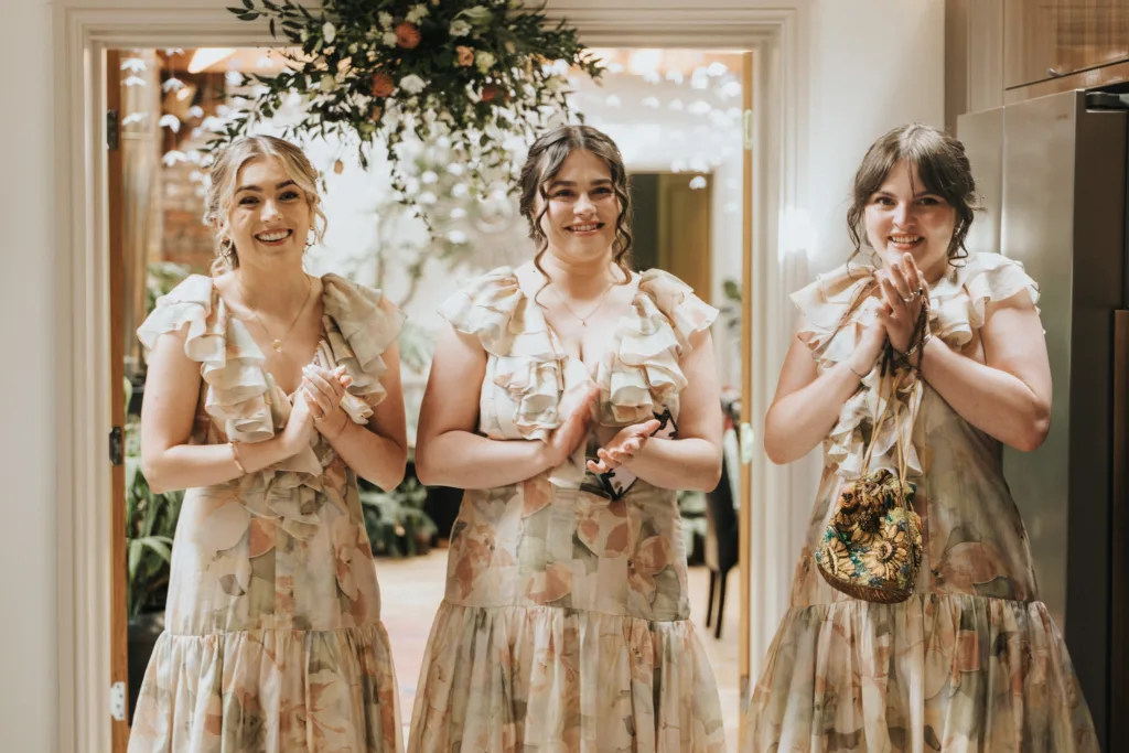 Three women stand side by side indoors, smiling and clapping. They are wearing matching floral dresses with frills. A flower arrangement hangs above them, and greenery is visible in the background. © Aimee Lince Photography, Wedding photographer in Lincolnshire