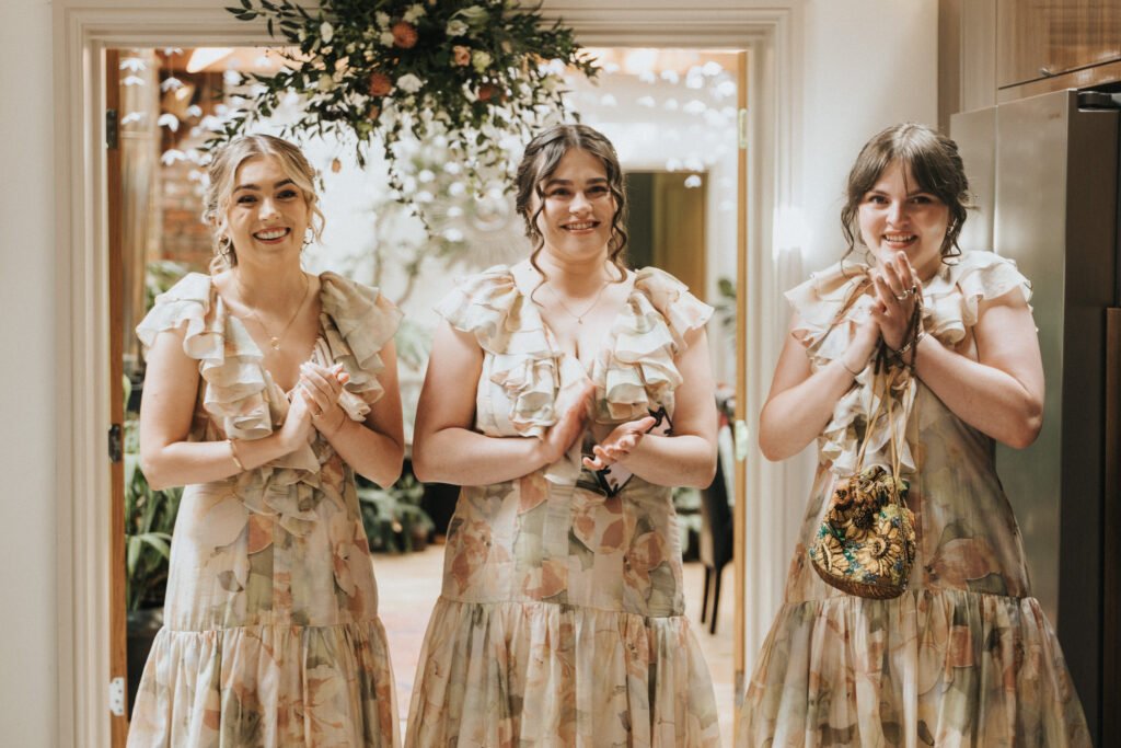 Three women stand side by side indoors, smiling and clapping. They are wearing matching floral dresses with frills. A flower arrangement hangs above them, and greenery is visible in the background. © Aimee Lince Photography, Wedding photographer in Lincolnshire
