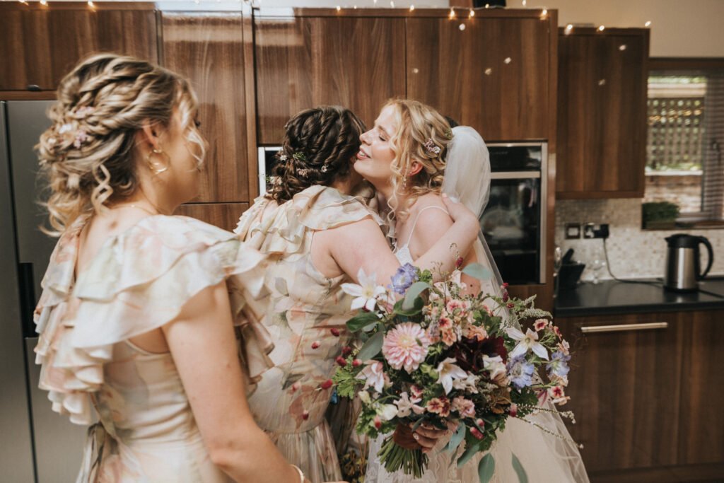 Three women in pastel floral dresses hug in a modern kitchen. One woman, wearing a veil and holding a colourful bouquet of flowers, smiles joyfully as the others embrace her. © Aimee Lince Photography, Wedding photographer in Lincolnshire