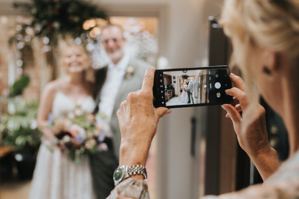 A woman takes a photo on her mobile of a smiling couple in wedding attire, standing close together and holding a bouquet, with the mobile capturing their image clearly. © Aimee Lince Photography, Wedding photographer in Lincolnshire