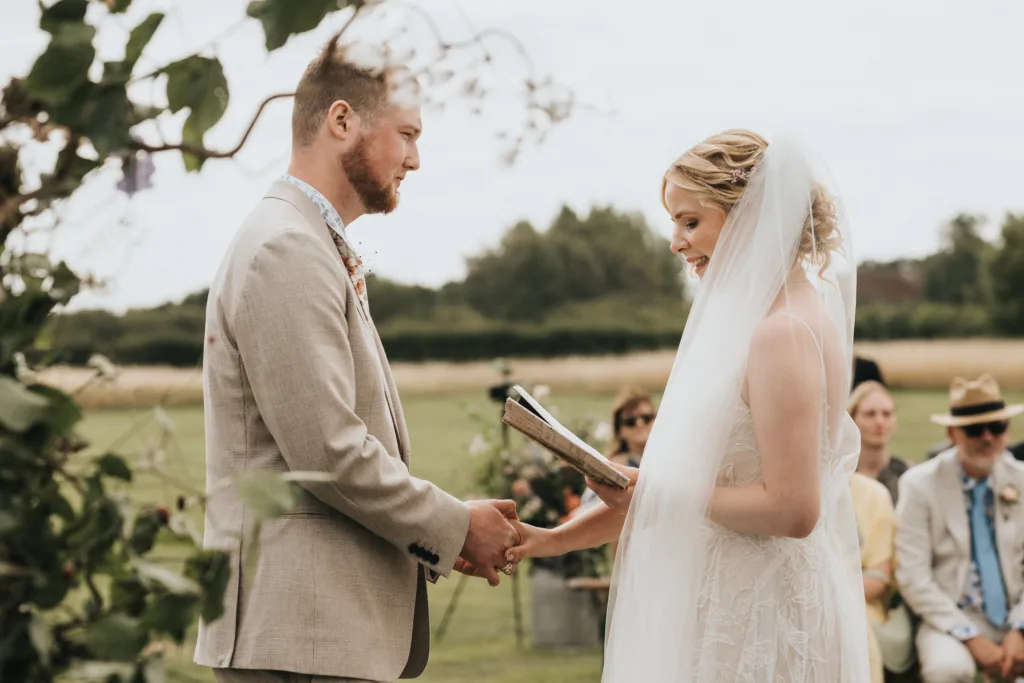 A bride and groom stand outdoors, holding hands and smiling at each other during their wedding ceremony, with guests seated in the background and greenery around them. © Aimee Lince Photography, Wedding photographer in Lincolnshire