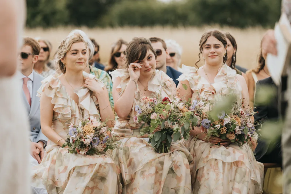 Three bridesmaids in matching floral dresses sit side by side, holding bouquets. The middle bridesmaid wipes away a tear, whilst the others smile softly. Guests in formal attire are visible in the blurred background. © Aimee Lince Photography, Wedding photographer in Lincolnshire