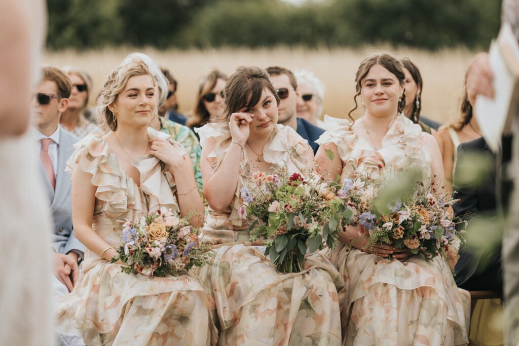 Three bridesmaids in matching floral dresses sit side by side, holding bouquets. The middle bridesmaid wipes away a tear, whilst the others smile softly. Guests in formal attire are visible in the blurred background. © Aimee Lince Photography, Wedding photographer in Lincolnshire