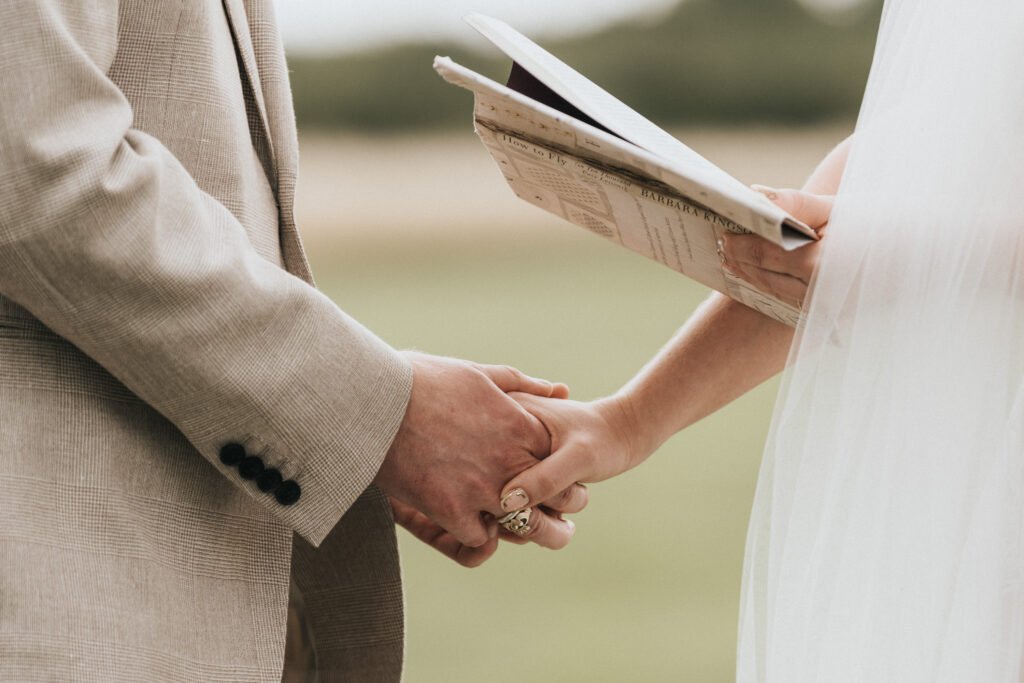 A close-up of two people holding hands outdoors, one wearing a beige suit and the other in white, holding papers, possibly during a wedding ceremony. The background is softly blurred greenery. © Aimee Lince Photography, Wedding photographer in Lincolnshire