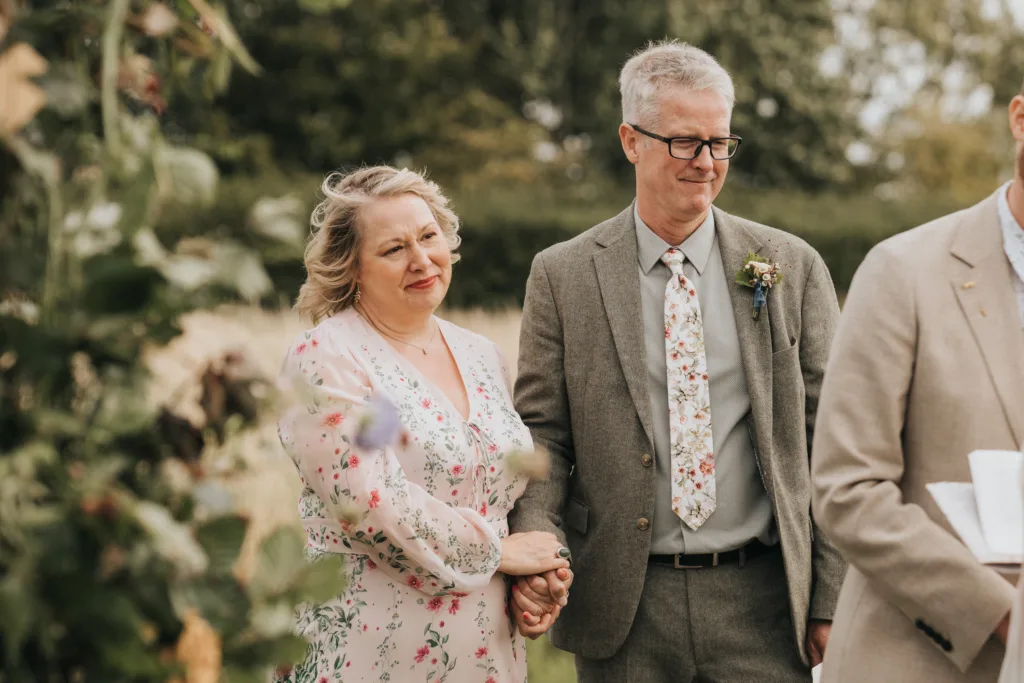A woman and man stand together outdoors, both dressed in floral-patterned clothing. The woman holds the man's arm and smiles softly, while the man wears glasses and a suit. Greenery and blurred trees are visible in the background. © Aimee Lince Photography, Wedding photographer in Lincolnshire