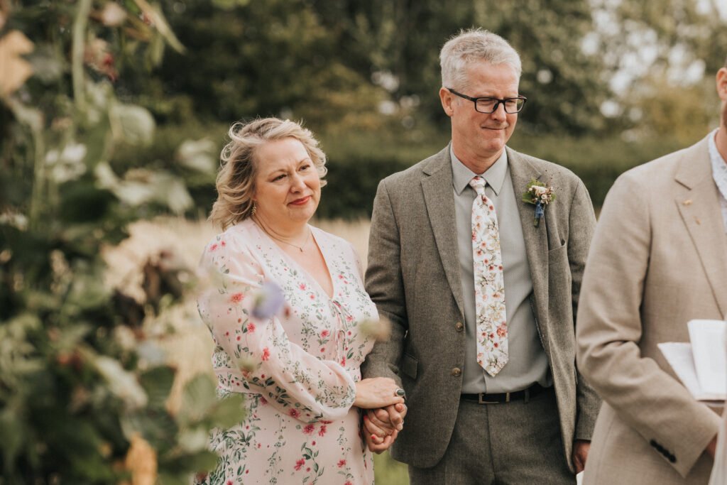 A woman and man stand together outdoors, both dressed in floral-patterned clothing. The woman holds the man's arm and smiles softly, while the man wears glasses and a suit. Greenery and blurred trees are visible in the background. © Aimee Lince Photography, Wedding photographer in Lincolnshire
