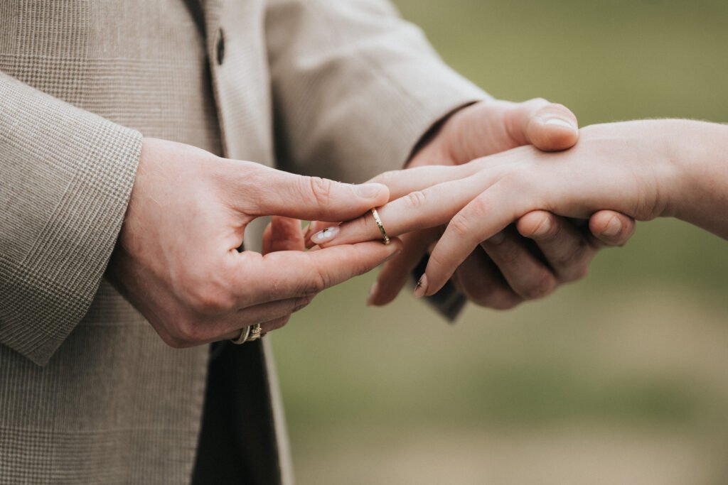 Close-up of a person in a beige suit gently placing a gold ring on another person's finger, symbolising a marriage or engagement ceremony, with a blurred green background. © Aimee Lince Photography, Wedding photographer in Lincolnshire