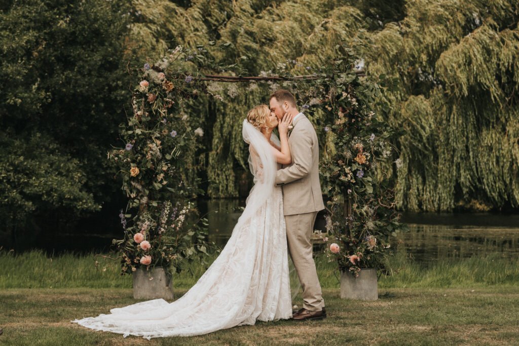 A bride and groom stand closely, touching faces and smiling, beneath a floral wedding arch outdoors, with lush green trees and a pond in the background. © Aimee Lince Photography, Wedding photographer in Lincolnshire