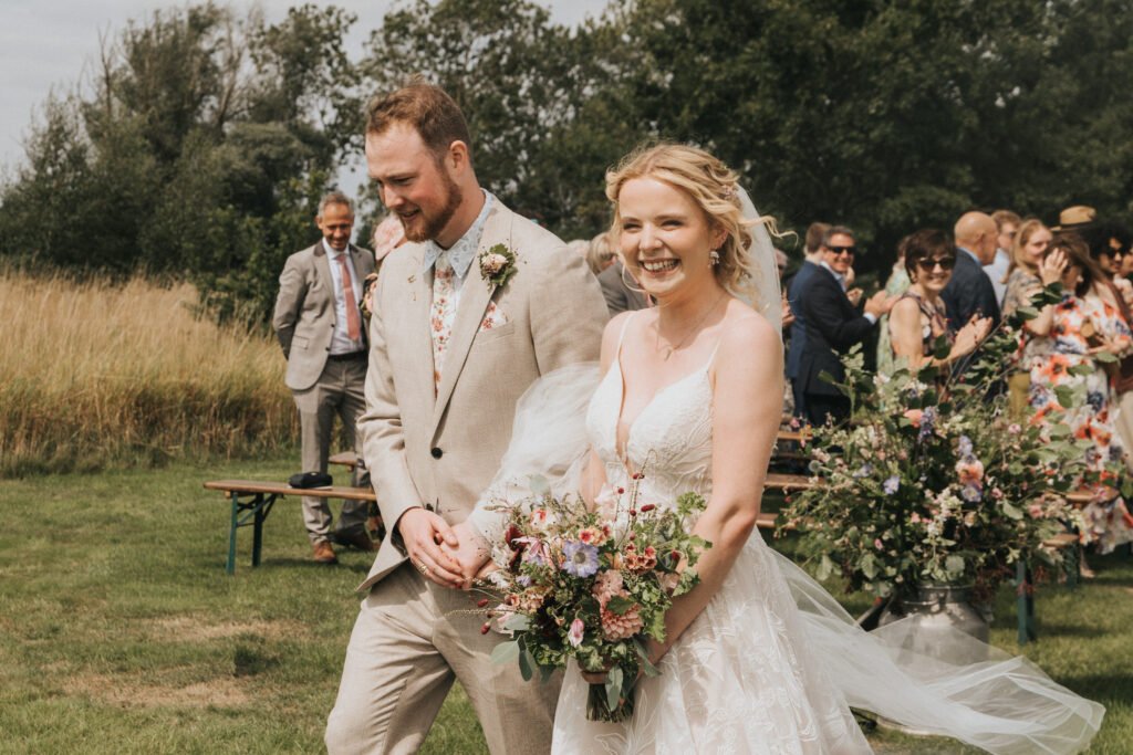 A bride and groom smile as they walk outdoors after their wedding ceremony, surrounded by guests and lush greenery. The bride holds a bouquet of flowers and wears a white dress; the groom wears a light tan suit. © Aimee Lince Photography, Wedding photographer in Lincolnshire