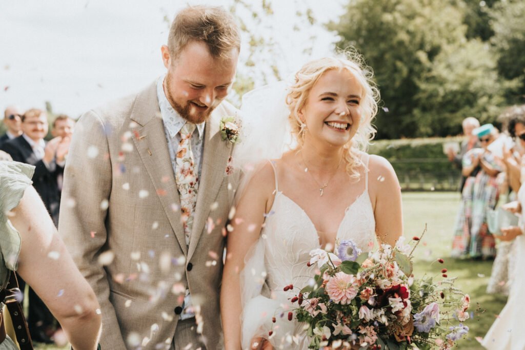 A joyful bride and groom walk outdoors, smiling as guests throw confetti. The bride holds a colourful bouquet and wears a white dress; the groom wears a light suit. Greenery and guests are visible in the background. © Aimee Lince Photography, Wedding photographer in Lincolnshire
