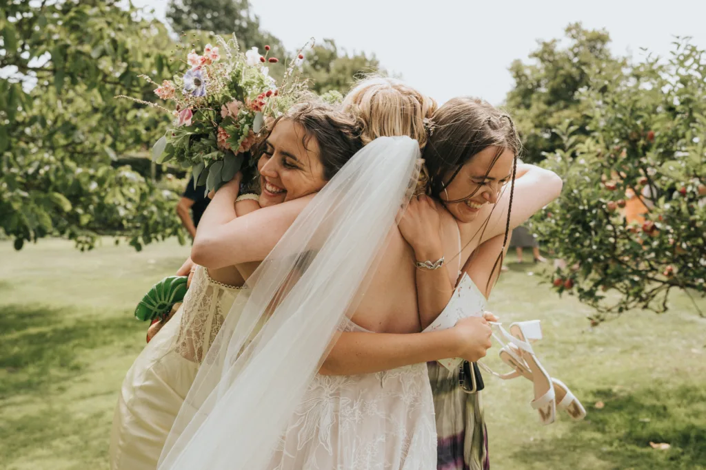 Three women, one in a wedding dress and veil holding a bouquet, share a joyful group hug outdoors in a green, sunny garden. All are smiling and appear happy, celebrating a special moment together. © Aimee Lince Photography, Wedding photographer in Lincolnshire
