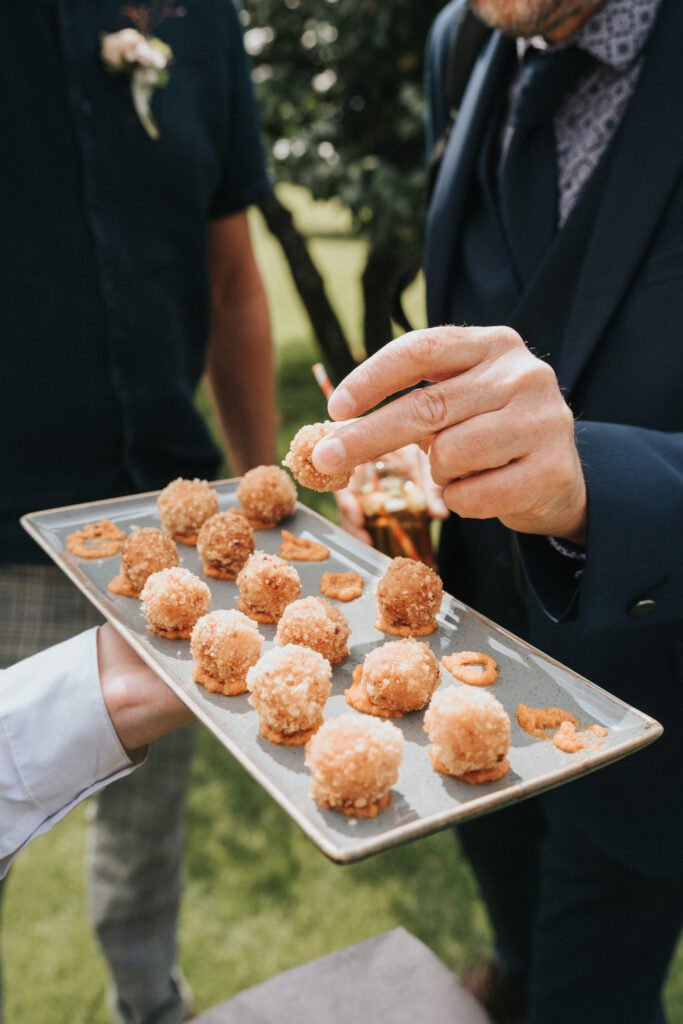 A person in a suit reaches to take a round, breaded starter from a rectangular tray being held by another person, with more starters and crispy rings on the tray. Outdoor setting with blurred greenery in the background. © Aimee Lince Photography, Wedding photographer in Lincolnshire