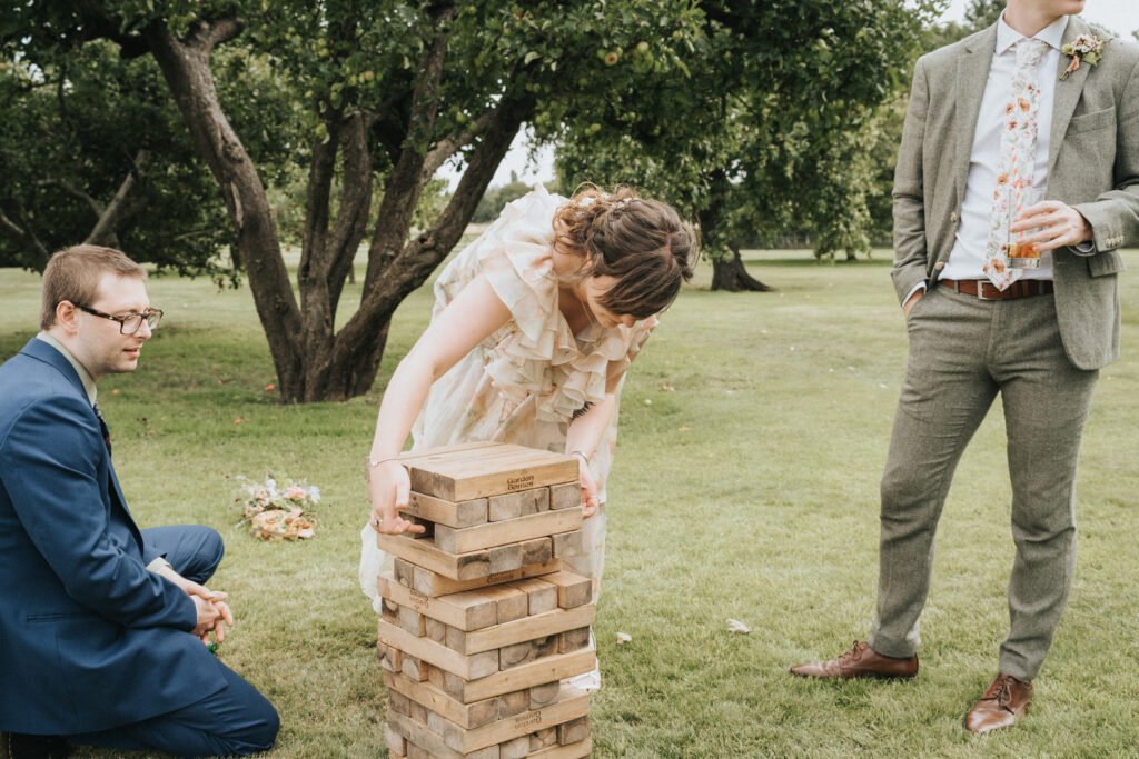 A woman in a floral dress carefully removes a block from a large outdoor Jenga tower on grass, while a man in a blue suit kneels nearby and another man in a grey suit stands holding a drink. Trees are in the background. © Aimee Lince Photography, Wedding photographer in Lincolnshire