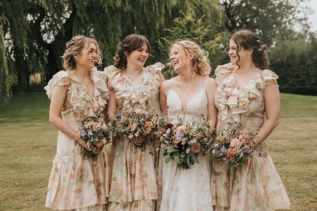 Four women stand together outdoors, smiling and holding bouquets. Three wear matching frilled dresses with floral patterns, while the fourth, in the centre, wears a white wedding dress. Lush greenery is in the background. © Aimee Lince Photography, Wedding photographer in Lincolnshire