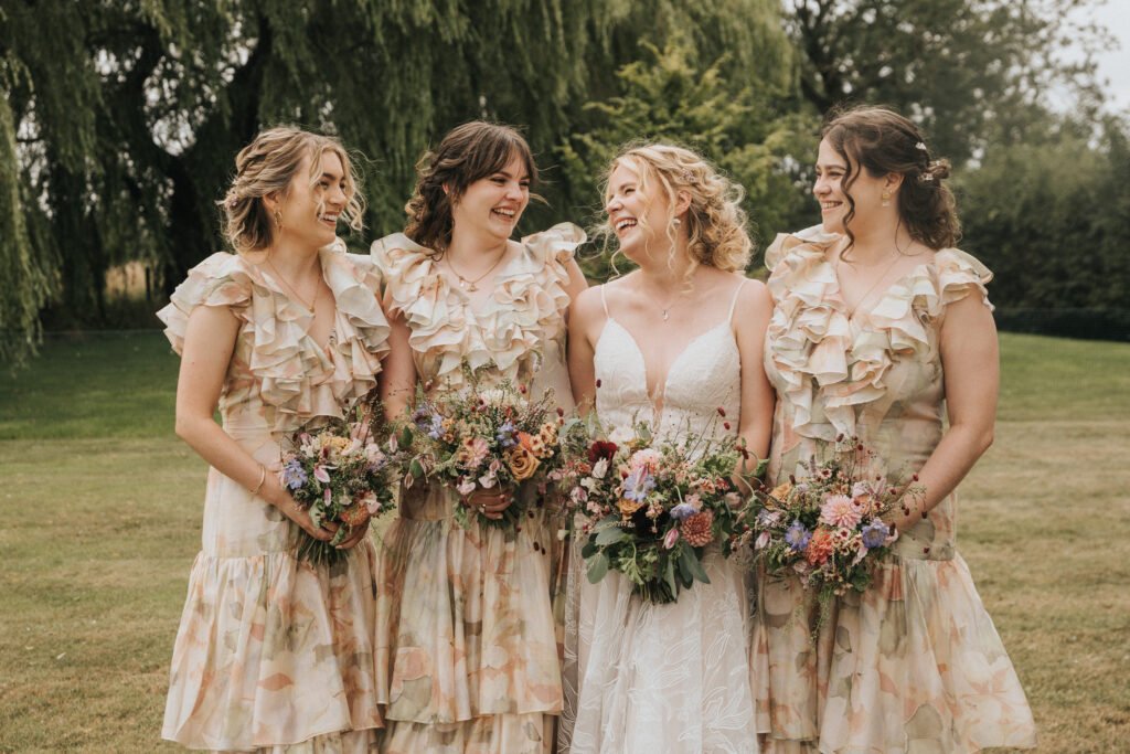 Four women stand together outdoors, smiling and holding bouquets. Three wear matching frilled dresses with floral patterns, while the fourth, in the centre, wears a white wedding dress. Lush greenery is in the background. © Aimee Lince Photography, Wedding photographer in Lincolnshire