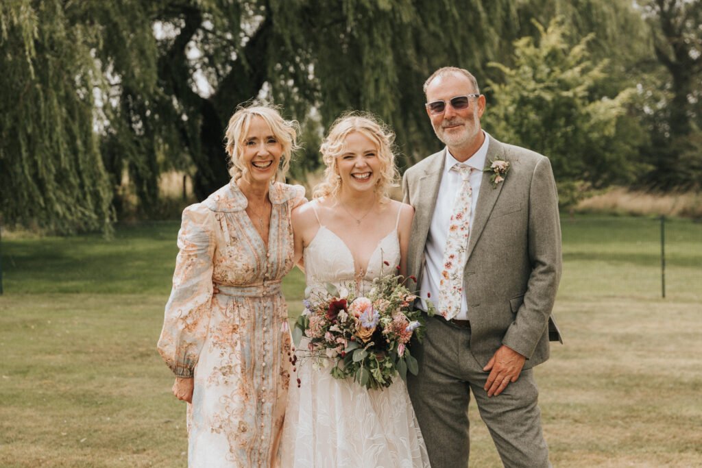 A bride in a white dress stands smiling outdoors, holding a bouquet, flanked by an older woman in a light floral dress and an older man in a grey suit with a floral tie; trees and grass are in the background. © Aimee Lince Photography, Wedding photographer in Lincolnshire