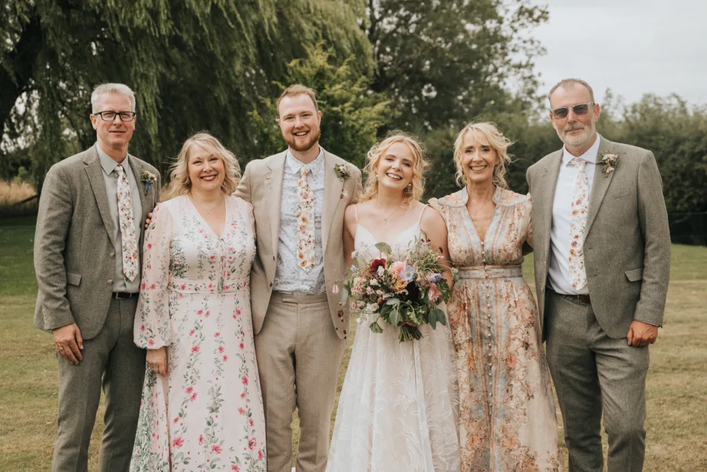 Six people stand outdoors on grass, smiling at the camera. The group includes three men in light suits and floral ties and three women in floral dresses. The central woman in white holds a bouquet, suggesting a wedding. Trees are in the background. © Aimee Lince Photography, Wedding photographer in Lincolnshire
