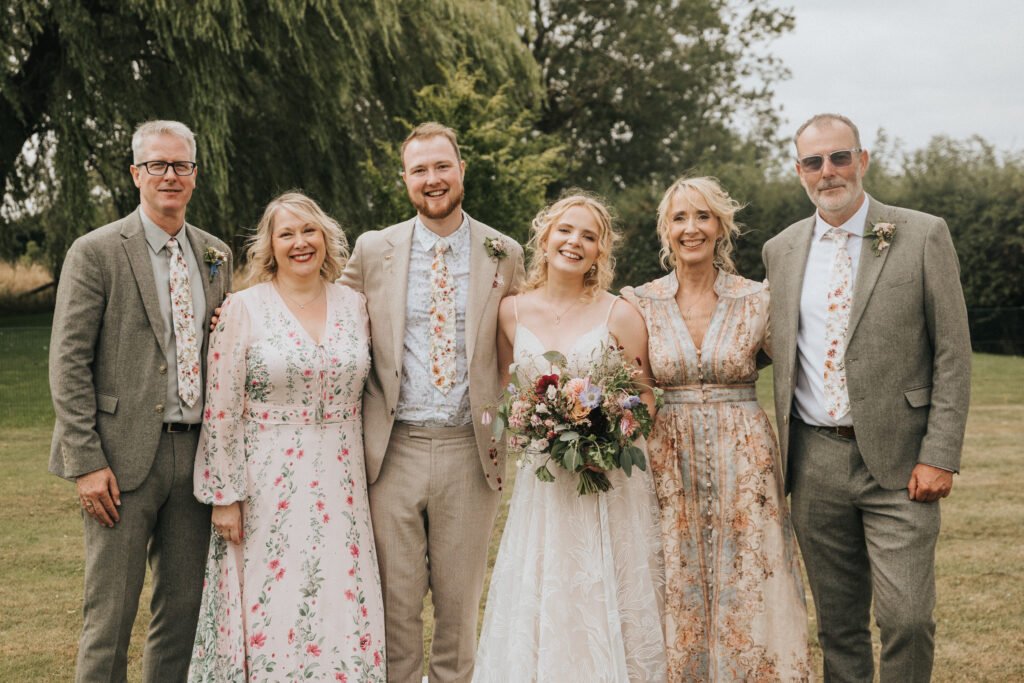Six people stand outdoors on grass, smiling at the camera. The group includes three men in light suits and floral ties and three women in floral dresses. The central woman in white holds a bouquet, suggesting a wedding. Trees are in the background. © Aimee Lince Photography, Wedding photographer in Lincolnshire
