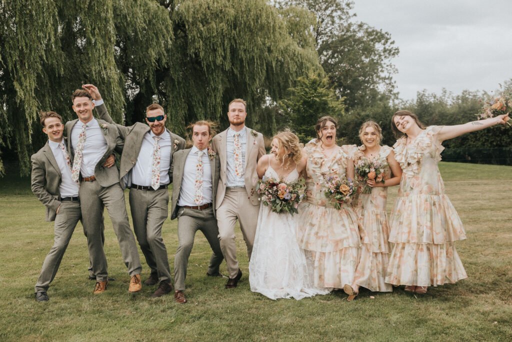 A wedding party poses playfully outdoors; the bride in white holds bouquets with bridesmaids in matching floral dresses, while groomsmen in grey suits and floral ties stand beside them under green trees. © Aimee Lince Photography, Wedding photographer in Lincolnshire