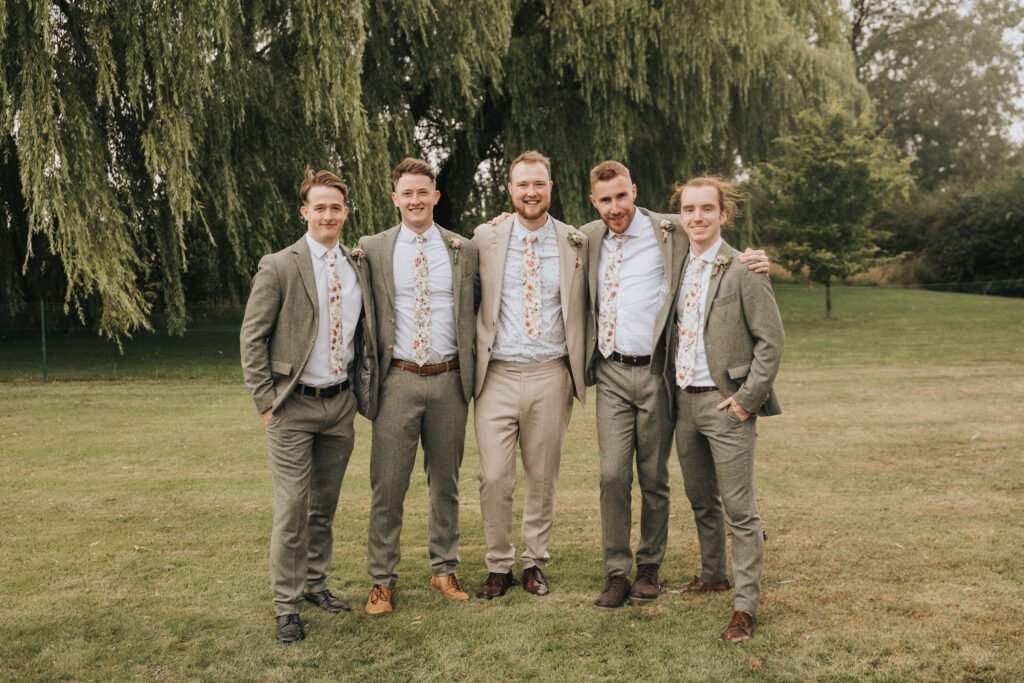 Five men stand together on grass in front of leafy trees, all wearing light-coloured suits and matching floral ties, smiling with arms round each other in a relaxed outdoor setting. © Aimee Lince Photography, Wedding photographer in Lincolnshire