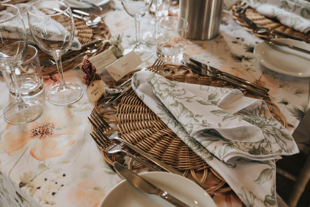 A close-up of an elegant table setting with woven placemats, floral serviettes, silver cutlery, wine glasses, and small name cards on a floral tablecloth. The atmosphere is refined and inviting. © Aimee Lince Photography, Wedding photographer in Lincolnshire