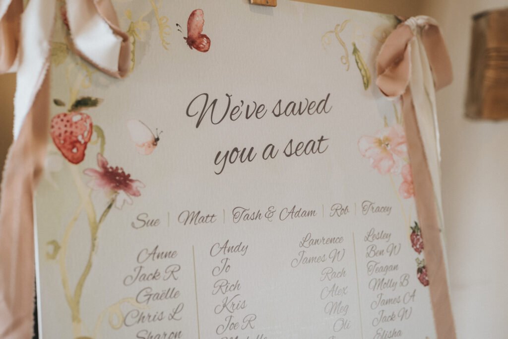A decorative seating plan with floral and butterfly illustrations, titled "We've saved you a seat," followed by a list of names written in elegant script. Pink ribbons are attached at the corners. © Aimee Lince Photography, Wedding photographer in Lincolnshire