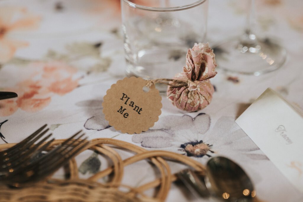 A small fabric pouch with a tag reading "Plant Me" sits on a floral tablecloth beside a drinking glass, cutlery, and a name card at an elegantly set table. © Aimee Lince Photography, Wedding photographer in Lincolnshire