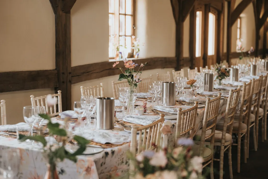 A long, elegant dining table is set for an event with floral arrangements, glassware, cutlery, and white chairs in a rustic room with wooden beams and natural light from windows. © Aimee Lince Photography, Wedding photographer in Lincolnshire