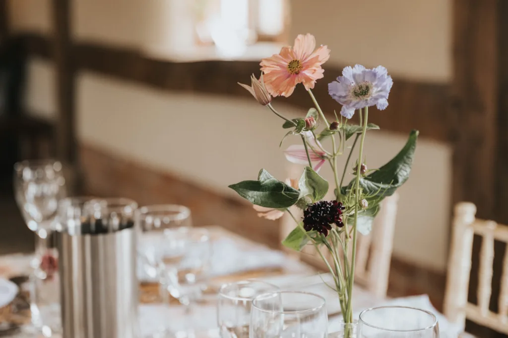 A close-up of a dining table set with wine glasses and cutlery, featuring a vase with pink, purple, and dark red flowers as the centrepiece. The background is softly blurred. © Aimee Lince Photography, Wedding photographer in Lincolnshire