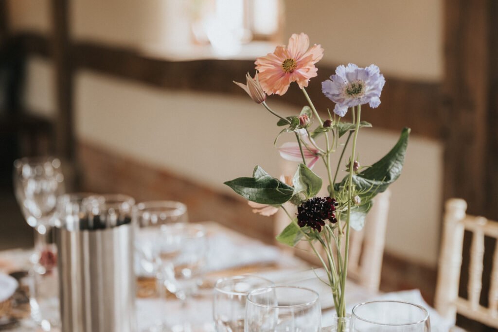A close-up of a dining table set with wine glasses and cutlery, featuring a vase with pink, purple, and dark red flowers as the centrepiece. The background is softly blurred. © Aimee Lince Photography, Wedding photographer in Lincolnshire