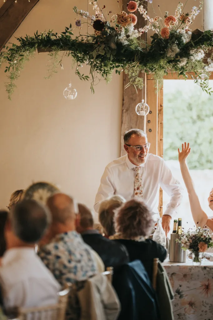 A man in a white shirt and floral tie stands smiling and speaking to a seated crowd at a wedding reception, with floral decorations and natural light coming through a window behind him. © Aimee Lince Photography, Wedding photographer in Lincolnshire