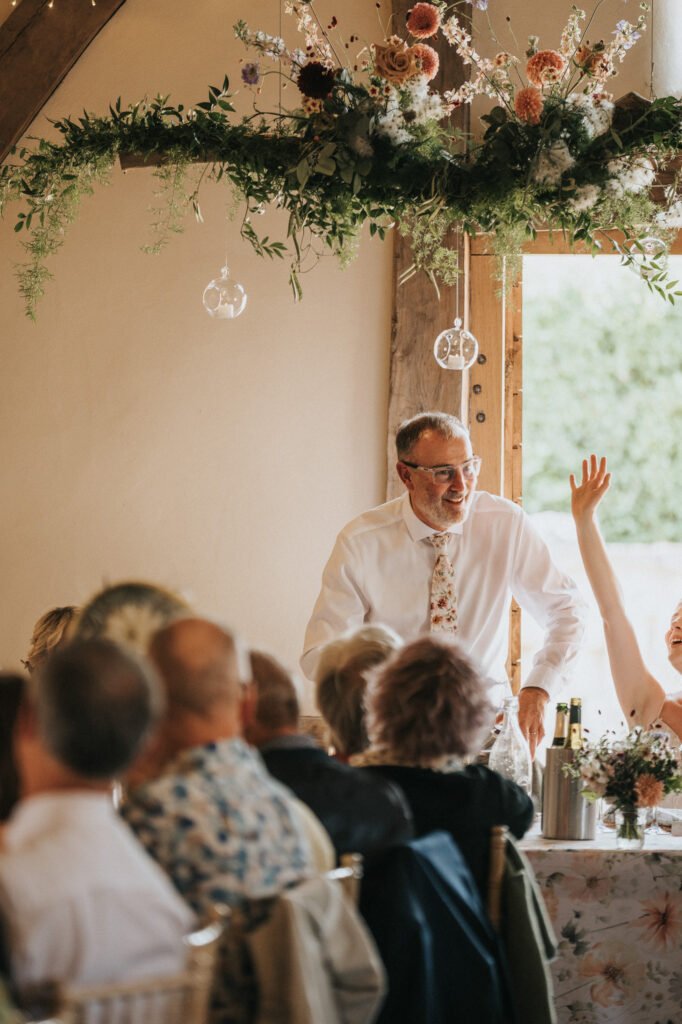 A man in a white shirt and floral tie stands smiling and speaking to a seated crowd at a wedding reception, with floral decorations and natural light coming through a window behind him. © Aimee Lince Photography, Wedding photographer in Lincolnshire