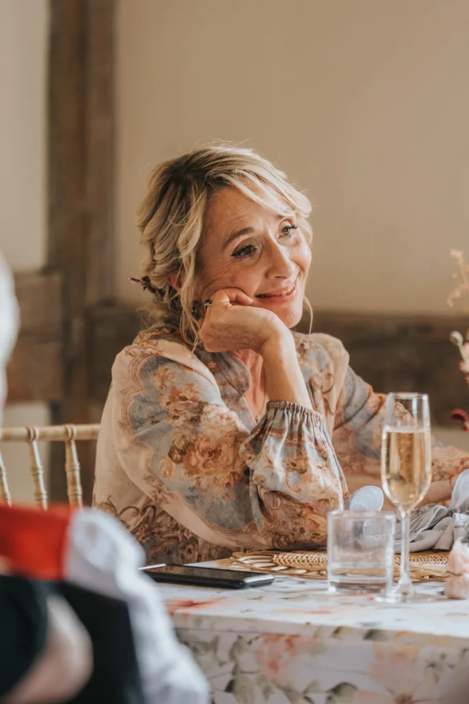A woman with blonde hair in an updo and wearing a floral blouse sits at a decorated table, resting her chin on her hand and smiling, with a glass of champagne in front of her. © Aimee Lince Photography, Wedding photographer in Lincolnshire