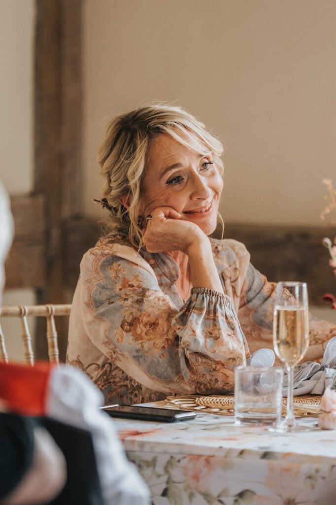 A woman with blonde hair in an updo and wearing a floral blouse sits at a decorated table, resting her chin on her hand and smiling, with a glass of champagne in front of her. © Aimee Lince Photography, Wedding photographer in Lincolnshire