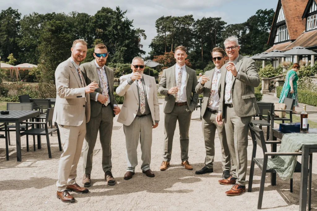 Six men in light suits and floral ties stand outdoors, smiling and holding drinks. They are gathered on a patio with empty tables and chairs, with greenery and a large building in the background on a sunny day. © Aimee Lince Photography, Wedding photographer in Lincolnshire